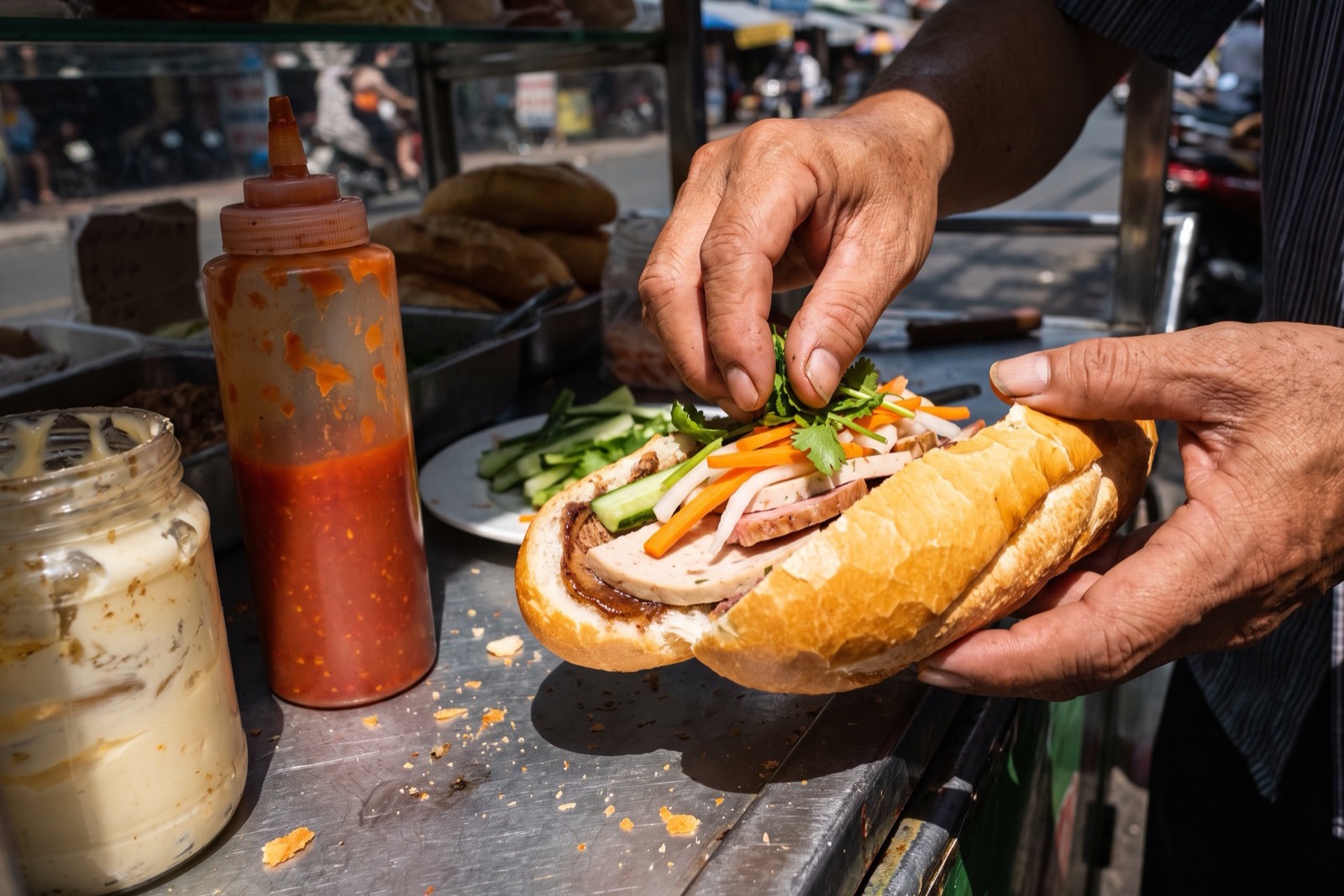 Vietnamese banh mi sandwich being assembled at a street cart with pate, cold cuts, pickled vegetables, and fresh herbs.